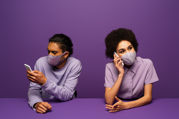 joven pareja afroamericana en máscaras protectoras usando teléfonos inteligentes aislados en púrpura  - Foto, Imagen