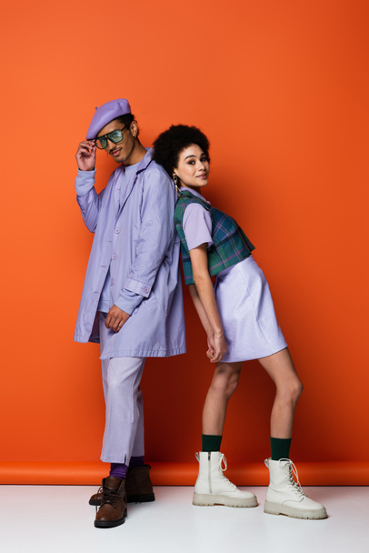 full length of trendy african american woman smiling while leaning on stylish man on orange  - Foto, Imagen