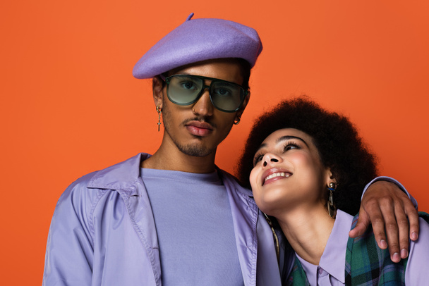 trendy uomo afroamericano in berretto e occhiali da sole abbracciando donna felice isolato su arancione  - Foto, immagini