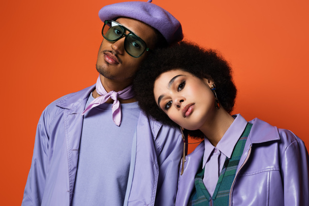 stylish woman in purple jacket leaning on man in beret and sunglasses isolated on orange - Photo, Image