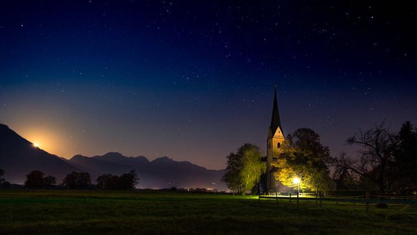 Moonlight shining from behind the Alps on small bavarian village church of Nussdorf am Inn by night - Foto, imagen
