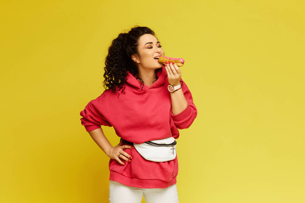 Young woman in a pink hoodie eating delicious eclair over the yellow background - Foto, immagini