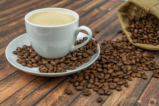 A cup of creamy coffee with roasted coffee beans next to it, wooden background - Foto, Imagen