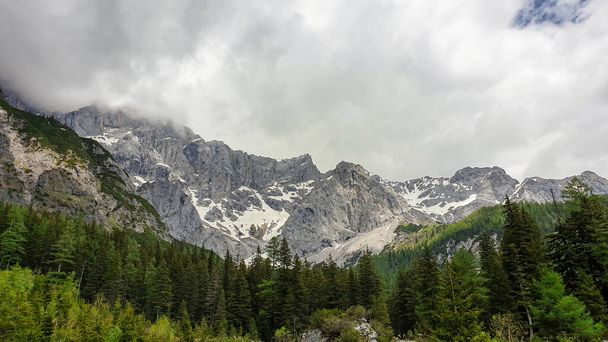 A panoramic view on the Alpine peaks in Austria from the Marstein. The slopes are still partially covered with snow. Stony and sharp mountains. Overcast. Baren slopes, dense forest at the foothill. - Valokuva, kuva