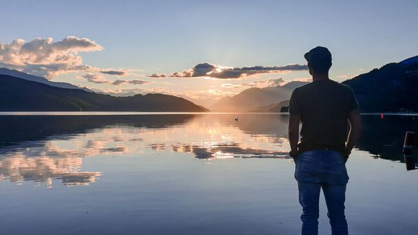 A man in a full cup standing at the shore of Millstaettersee and enjoying the sunset. The sun sets behind high Alps. Calm surface of the lake reflects the orange sky and the mountains. Meditation - Photo, Image