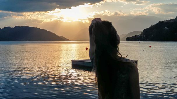 Mavi elbiseli bir kadın Millstaettersee Gölü kıyısında uzun, kahverengi saçlarını savuruyor. Güneş yüksek Alpler 'in arkasında batıyor. Gölün sakin yüzeyi turuncu gökyüzünü ve dağları yansıtır. Eğlenceli. - Fotoğraf, Görsel