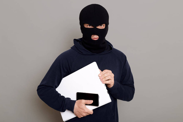 Satisfied man robber steals laptop beech and phone, being dressed in robber mask and black turtleneck, looks at camera firmly holds stolen things, posing isolated over gray background. - Zdjęcie, obraz