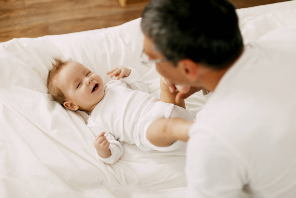 Boldog apa játszik imádnivaló baba a hálószobában egy hófehér ágy - Fotó, kép