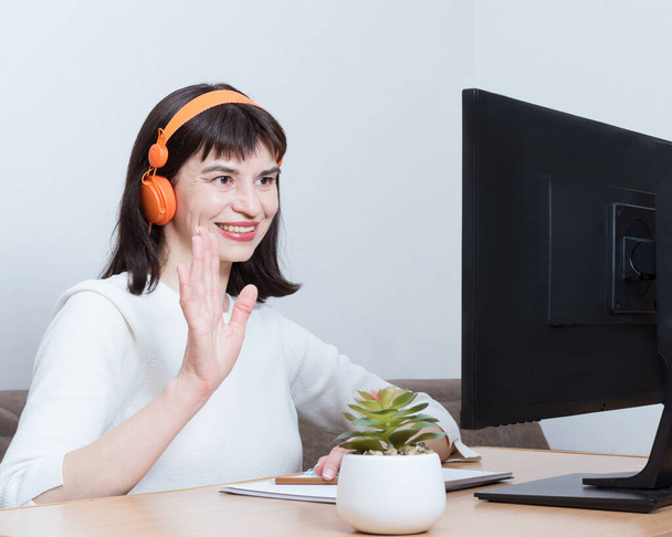 Чарівна жінка в помаранчевих навушниках з відеодзвінком і вітальний жест перед монітором комп'ютера
. - Фото, зображення