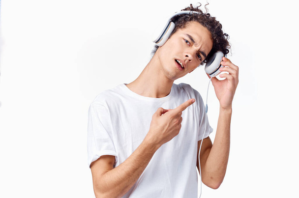 šťastný kudrnatý chlapík ve sluchátkách a v bílém tričku poslouchá hudbu a drží si ruce u obličeje - Fotografie, Obrázek
