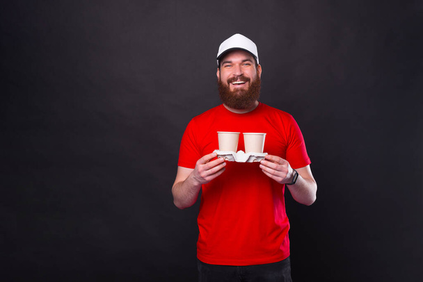 Young bearded hipster smiles at the camera while holding two paper takeaway cups over black background in a studio. - Photo, Image