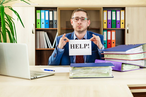 Boss businessman at his desk in the office with a bunch of folders with papers. Holds a HELP sign in his hands. Strenuous office work manager. Preparing of report by an office worker - Photo, Image