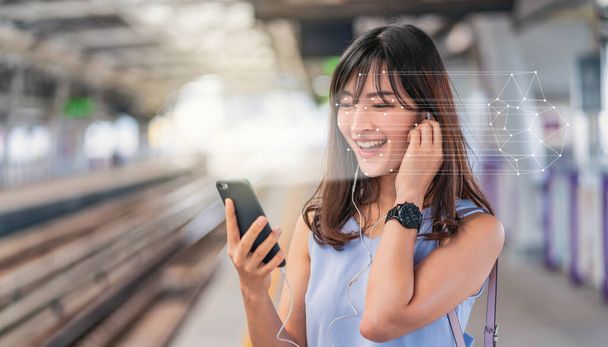 Ασιάτισσα που χρησιμοποιεί αναγνώριση προσώπου μέσω έξυπνου κινητού τηλεφώνου και ακούει μουσική στην Πλατφόρμα Σιδηροδρόμων, Βιομετρική Επαλήθευση και τεχνητή νοημοσύνη - Φωτογραφία, εικόνα