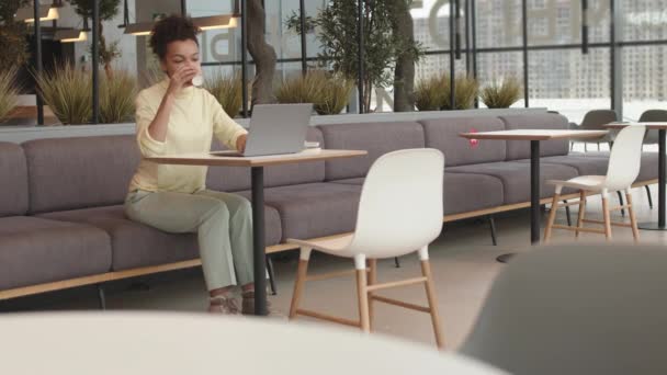 Pełne ujęcie młodej mieszanej kobiety siedzącej samotnie przy stole w nowoczesnej przestrzeni dla współpracowników, pijącej z papierowej filiżanki, korzystającej z przenośnego komputera - Materiał filmowy, wideo