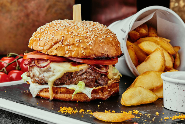 Big burger with juicy cutlet, cheese, tomato, lettuce, served with french fries and barbecue sauce on a black plate on the background of copper plates. close up - Foto, imagen
