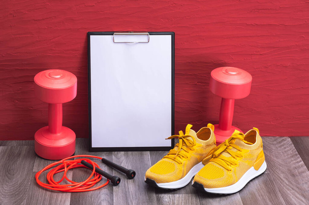 exercícios em casa - halteres vermelhos, corda de salto e tênis amarelos, quadro de notas, close-up e fundo vermelho - Foto, Imagem