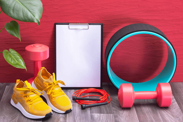 training at home - red dumbbells, jump rope and yellow sneakers, note board, massage roller and green large leaves on a red background - Zdjęcie, obraz