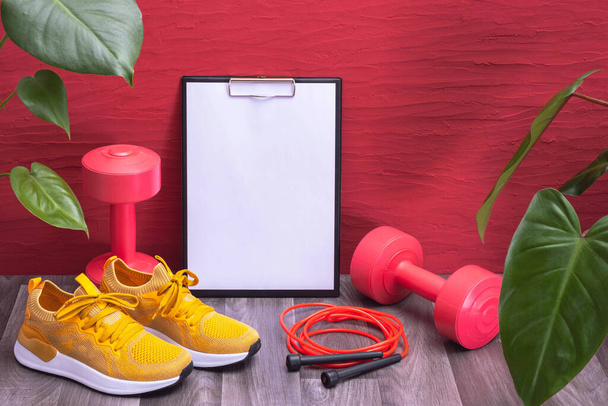 home cvičení - červené činky, švihadlo a žluté tenisky, poznámkový blok a zelené velké listy na červeném pozadí, detailní záběr - Fotografie, Obrázek