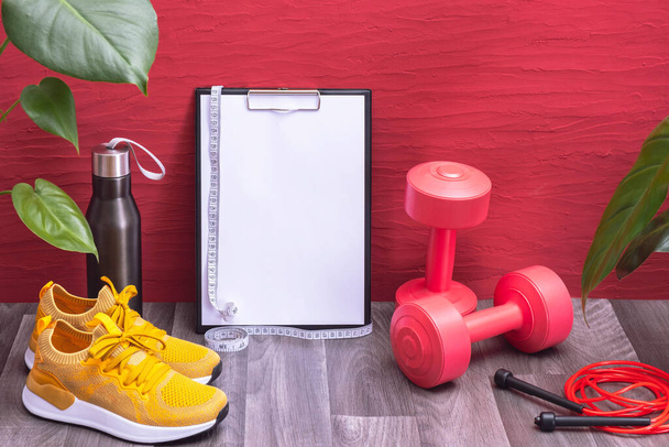 workout at home - red dumbbells, water bottle and jump rope, note board and measurement tape, yellow sneakers and big green leaves, red background - Fotoğraf, Görsel