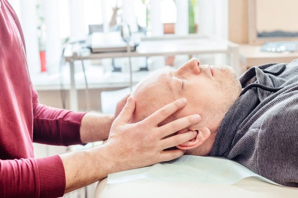 Mężczyzna poddawany terapii czaszkowej, leżący na stole do masażu w gabinecie leczenia osteopatycznego CST, osteopatii i terapii manualnej - Zdjęcie, obraz