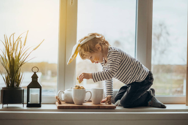 Cute little boy toddler with bunny ears headband sitting on big cozy and pretty decorated windowsill, l oking at window, enjoying view outdoor, feeling comfort with reliable window systems - Photo, Image