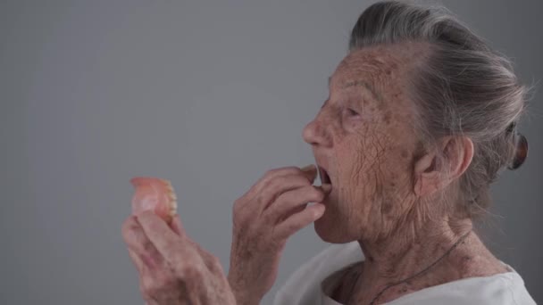 Mujer mayor caucásica muy vieja de 90 años con arrugas profundas y pelo gris ajustado prótesis dental sobre fondo de pared gris en el estudio. Mujer mayor inserta prótesis dentales. Dientes falsos. Concepto dental - Metraje, vídeo