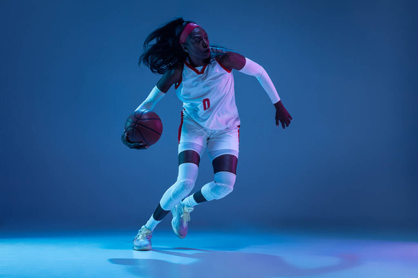 Beautiful african-american female basketball player in motion and action in neon light on blue background. Concept of healthy lifestyle, professional sport, hobby. - Фото, изображение