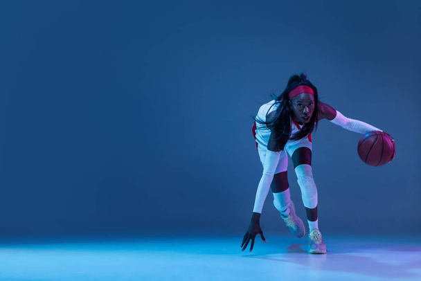 Beautiful african-american female basketball player in motion and action in neon light on blue background. Concept of healthy lifestyle, professional sport, hobby. - Foto, afbeelding