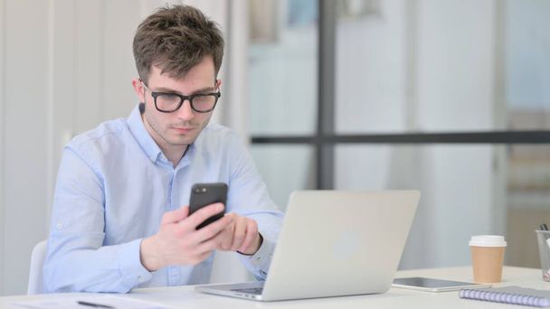 Молодой человек с ноутбуком с помощью смартфона на работе - Фото, изображение