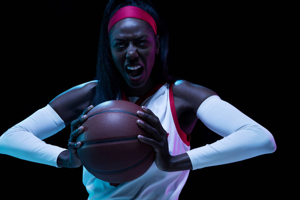 Beautiful african-american female basketball player in motion and action in neon light on black background. Concept of healthy lifestyle, professional sport, hobby. - Photo, image