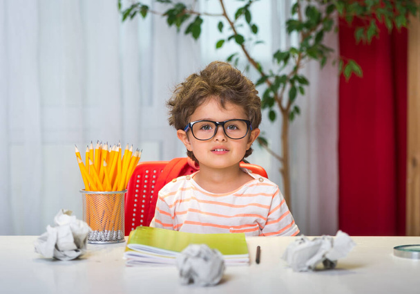 Портрет маленького хлопчика, який дивиться на камеру з посмішкою. Малюк в окулярах з олівцем робить домашнє завдання вдома. Малюк початкового класу. Повернутися до школи. Нові норми
. - Фото, зображення