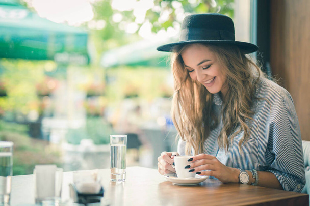 A cheerful beautiful Bosnian Caucasian woman in a hat holding a cup of coffee in a cafe - Foto, Bild