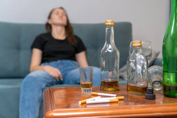 Opilá tmavovlasá žena sedící na gauči, soustředěná na prázdné láhve, cigarety. Sociální problém alkoholismu - Fotografie, Obrázek