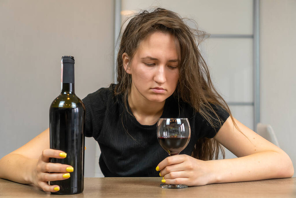 Молодая красивая темноволосая женщина с взъерошенными волосами в депрессии, расстроенные чувства, апатия пьет алкоголь за столом - Фото, изображение