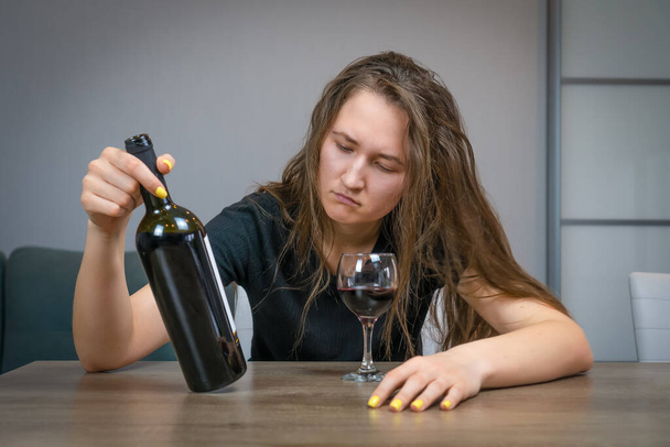 Jeune belle femme brune aux cheveux touffus dans la dépression, sentiments contrariés, apathie regarde une bouteille de vin à la table - Photo, image