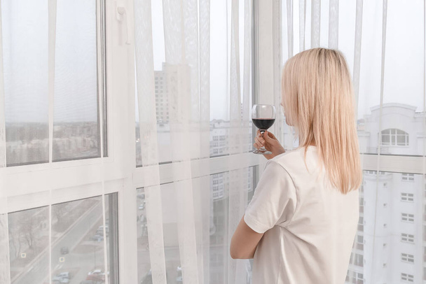 Nuori vaalea nainen valkoisessa T-paidassa punaviinilasillisen kanssa seisoo ikkunassa ja katsoo kaupungin kaukaisuuteen. Suuret ikkunat makuuhuoneessa. Kuva takaa. - Valokuva, kuva