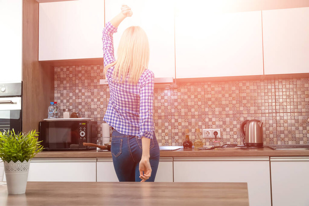 Allegro giovane donna bionda snella in jeans blu e una camicia cuochi in cucina e balli, spazio copia. Foto dal retro Foto con abbagliamento - Foto, immagini