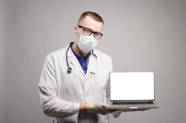mannelijke arts in een jas, masker, bril, handschoenen en met een stethoscoop met een laptop in zijn handen, geïsoleerd scherm, studio schieten - Foto, afbeelding