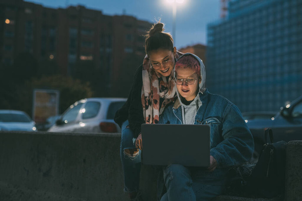 Dos mujeres caucásicas jóvenes al aire libre por la noche usando la computadora sonriendo feliz - Foto, imagen