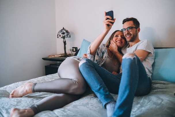 Young pregnant couple resting bed indoor at home using smartphone taking selfie - Foto, Imagem