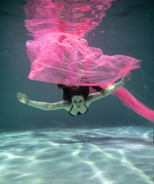 mooi meisje in een zwart badpak en met een roze pareo onderwater op een blauwe achtergrond - Foto, afbeelding