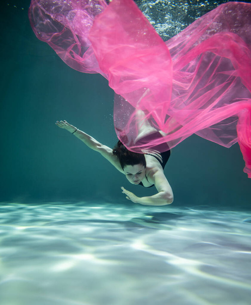 beautiful girl in a black swimsuit and with a pink pareo underwater on a blue background - Foto, Imagen