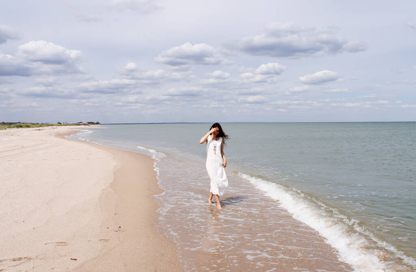Ελευθερία νεαρή γυναίκα φορώντας μακρύ λευκό φόρεμα περπατώντας στην παραλία - Φωτογραφία, εικόνα