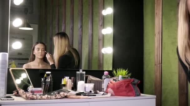 μακιγιάζ καλλιτέχνης κάνει μακιγιάζ για την ασιατική κορίτσι στο σαλόνι ομορφιάς, αντανάκλαση στον καθρέφτη - Πλάνα, βίντεο