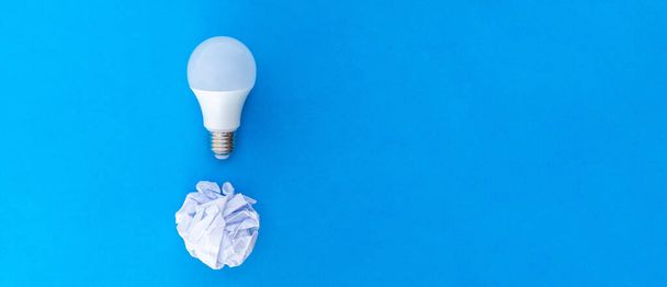 Ideia conceito representado por um papel branco amassado e uma lâmpada LED sobre um fundo azul e grande espaço de cópia. - Foto, Imagem