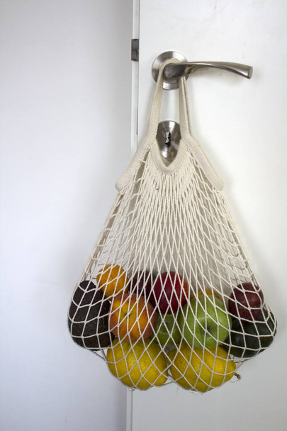 Mehrweg-Einkaufstasche mit Früchten am Türgriff. Umweltfreundliche Tasche aus Baumwolle. Gesundes Wohnkonzept.  - Foto, Bild