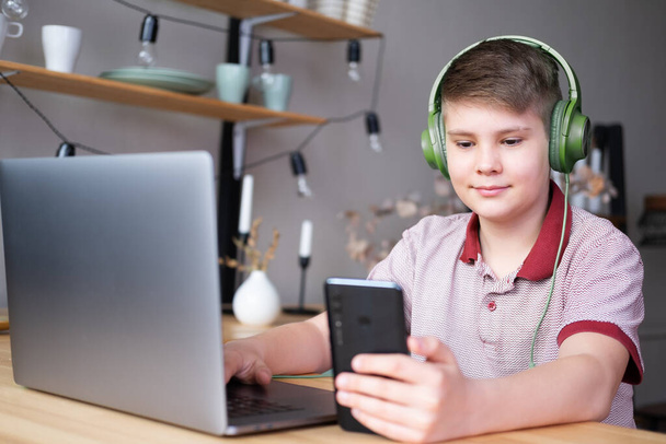 menino adolescente bonito em fones de ouvido estudando online, jogando em jogos de vídeo usando laptop e telefone. - Foto, Imagem