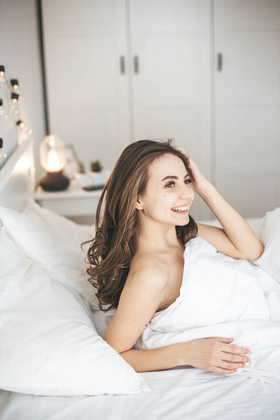 Beautiful young woman waking up in comfortable bed on white fresh sheets. - Photo, Image