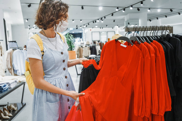 Vrouw gaat winkelen en koopt nieuwe kleren in een winkelcentrum met een gezichtsmasker tijdens een uitbraak en quarantaine van een coronavirus infectie - Foto, afbeelding