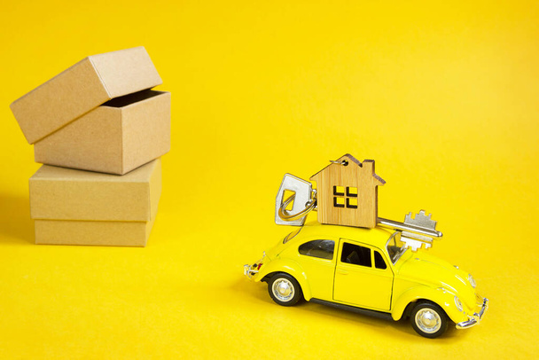 Yellow toy car with a key to the house on the roof on a illuminating background and packing boxes. Moving to a new home, mortgage, buying an apartment, taxi. Copy space. Kaluga, Russia, 08.04.21 - Fotoğraf, Görsel